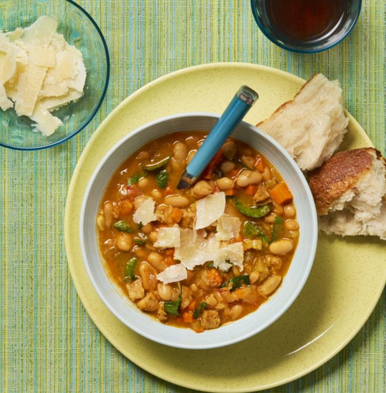 bowl of tuscan white bean soup on green background with a bowl of parmesan and a beverage